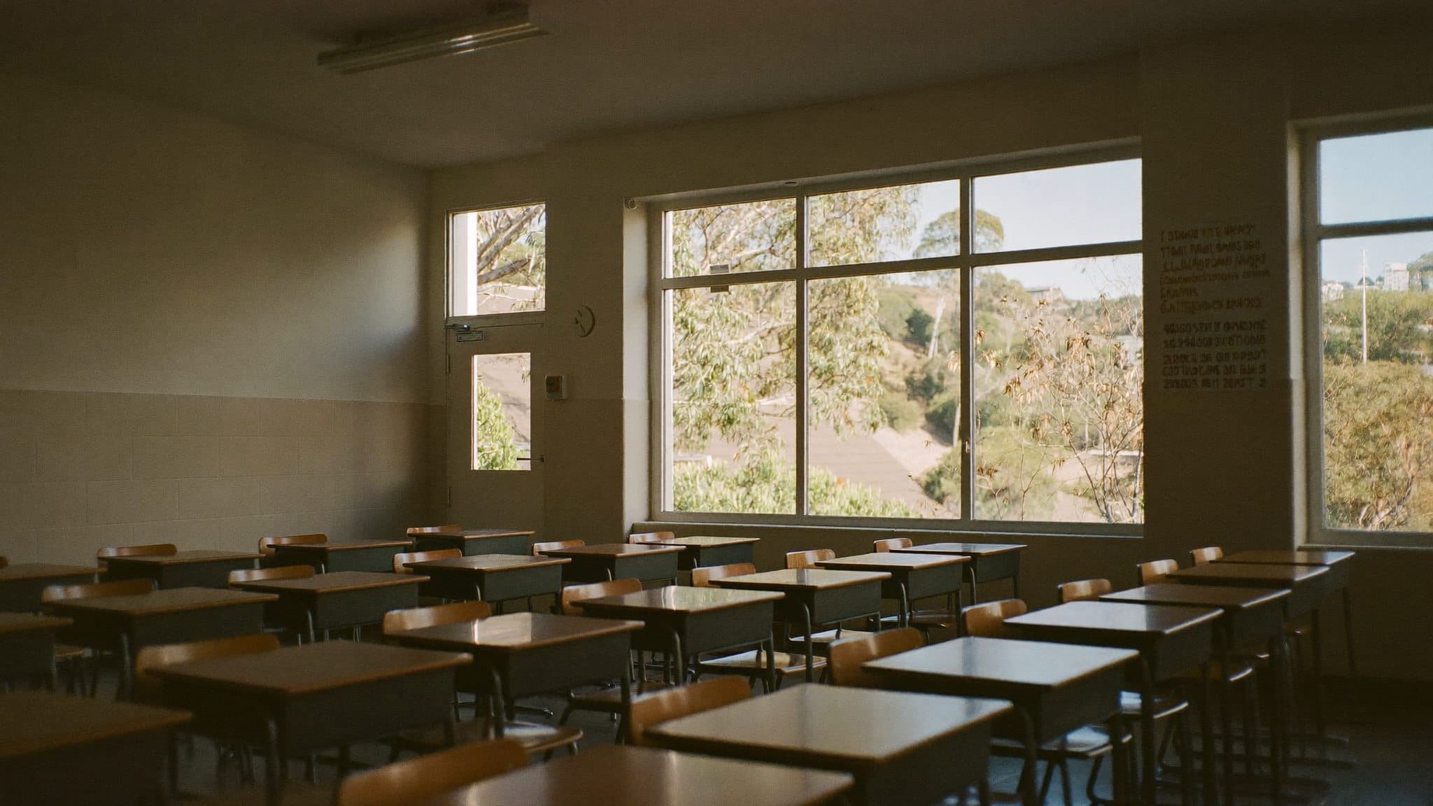 An empty Australian classroom early in the morning: sunlight across the desks, gum trees through the window.